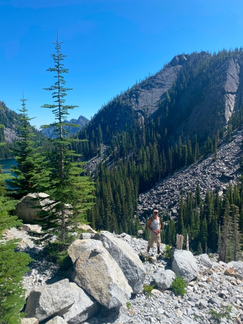 A man stands in a large landscape of mountains and tall pine trees and rocks with blue sky.