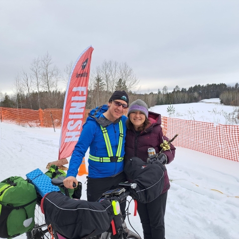 A man in a blue jacket and neon green harness holds his bag-laden bike and hugs his wife at a winter race finish line.