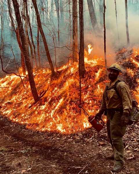 A firefighter smiles for the camera next to a hillside on fire with a drip torch in his hand