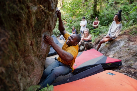 A man smiles while climbing a boulder. There are people in the background watching and smiling.