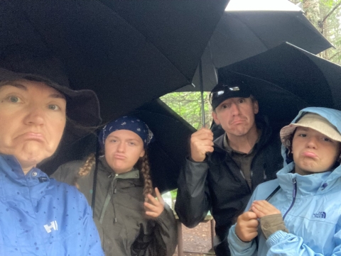 Four people stand under black umbrellas and make unhappy faces.