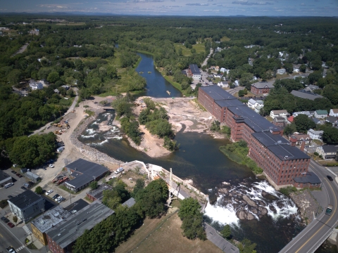 Aerial view of a river flowing through a city. Waterfalls, and a fishway channel are visible. Red brick buildings line the river bank. 