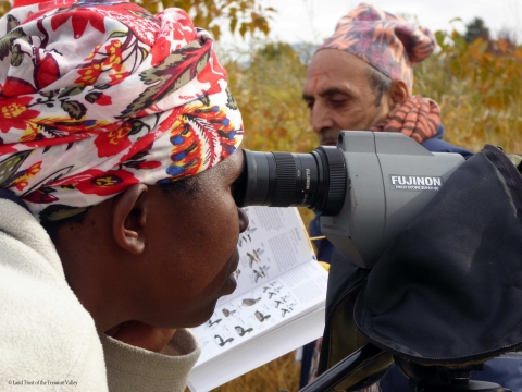 A woman looks through a spotting scope, and a man can be seen in the background behind the scope