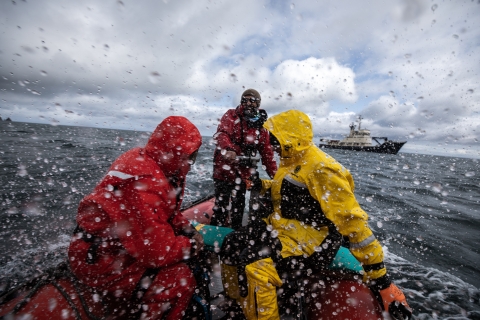 People in bright orange and yellow rain gear in an orange skiff boat through choppy waters through a rain spattered lens. A blue and white ship is in the background.