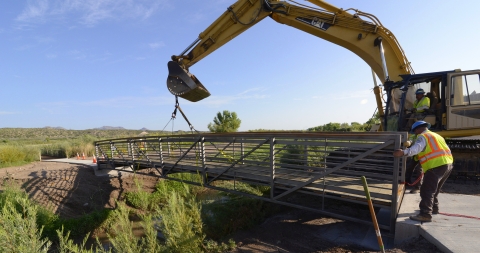 A crane operator lowers a bridge into place while a coworker on the ground adjusts the placement. 