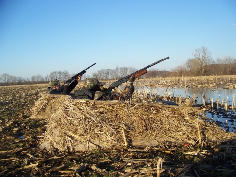 Two hunters in camouflage bunker with guns raised to the sky.