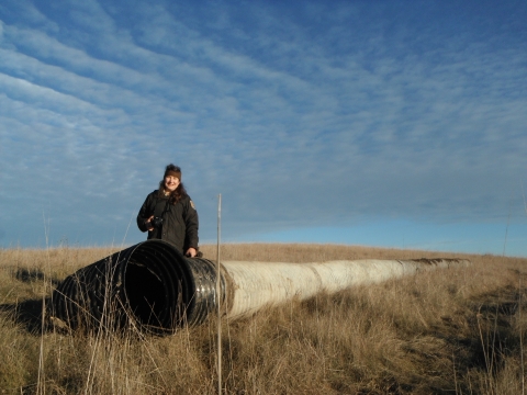 An uniformed adult stands in front of one end of a long drain pipe in a grassy prairie