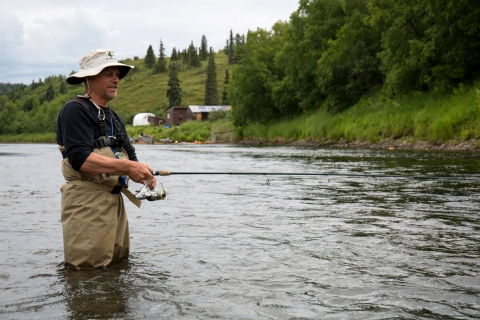 Man in fishing gear boots fishing in a river with hill of green grass and trees.