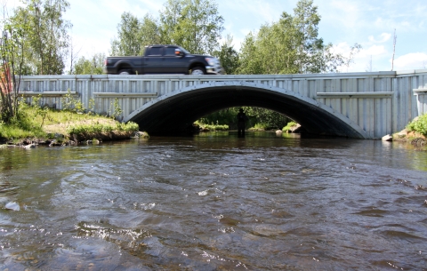 A black truck drives over a newly-constructed and much larger culvert or fish passage over a small river in Alaska.