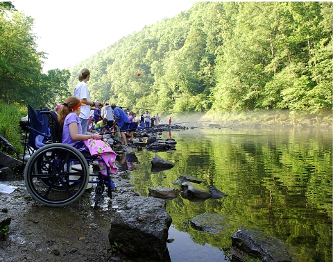 A line of children along a riverbank fishing, first one using wheelchair 