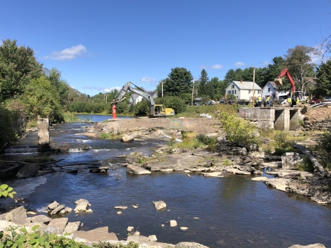 Concrete dam spans halfway across a river with heavy machinery and people in construction hats on top