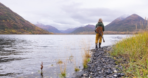 woman with a guitar stands on a lake shore looking out across the lake