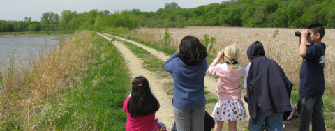 Five kids use binoculars to observe a goose outside on a gravel trail