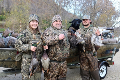 A woman and 2 men dressed in camouflage hold different species of ducks in front of their boat