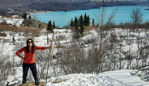 A woman standing wearing a bright orange shirt and sunglasses holding a walking stick in a snowy landscape. A bright, aqua blue lake in the background.