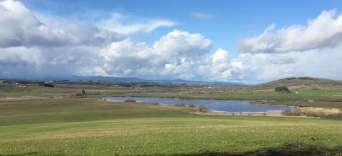 Wide view of prairie with pond in the distance on a partly cloudy day.