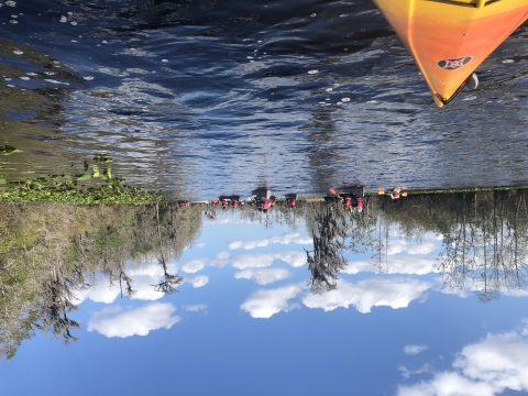 The bow of a kayak floats in the corner of a photo. Multiple canoes are seen in the background, filled with happy students paddling on the Okefenokee Swamp. 