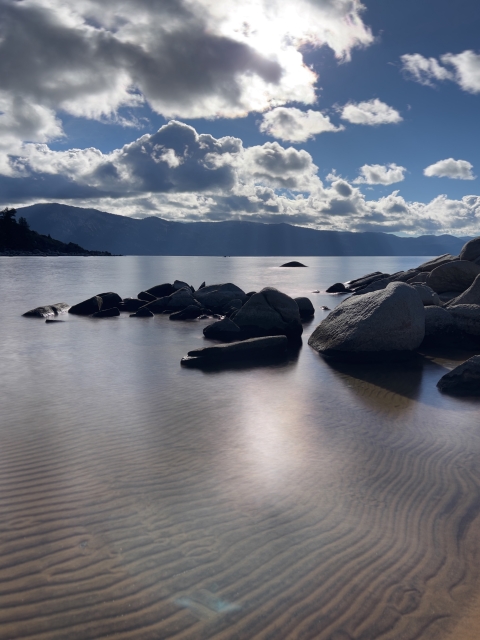 A lake with boulders in the forefront and mountains in the distance as clouds pass over the sun