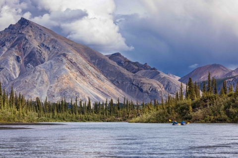 Paddlers navigate a stretch of river with firs along the banks and purple mountains rising behind them.