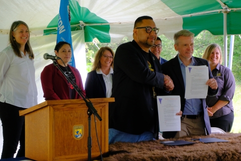 Five people stand behind two men who are shaking hands and hold up documents