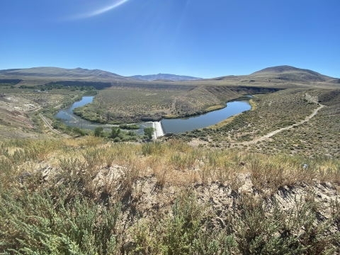 Image of a river flowing through a desert sage landscape with small mountains in the backdrop and bright blue skies.