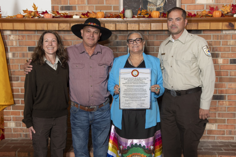 Four people stand together smiling as a woman in blue holds a signed document