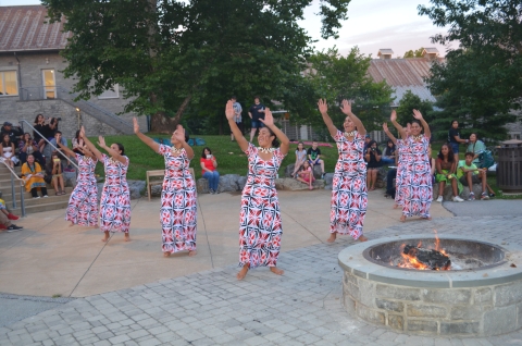  7 people in similar dresses with hands raised to left in unison dance outdoor amphitheater 