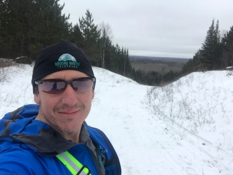 A man in a knit hat, sunglasses and a blue jacket smiles at the top of a snowy trail.