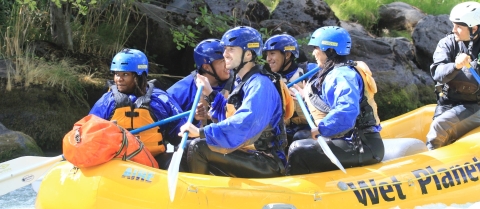 A group of people in yellow raft, rafting down a river. They are all wearing blue helmets and suits with orange life vests.