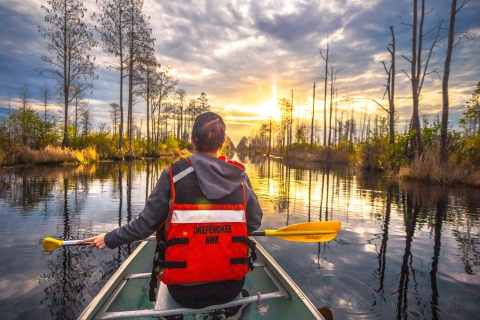 woman in front of canoe holding a yellow paddle in dark reflective water with cypress trees and clouds in a sunny sky. 