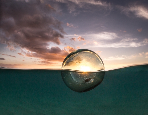 A glass ball floats in the ocean with the sun setting through it.