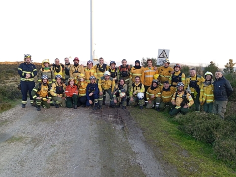 A large group of people in fire gear stand as a group and pose for a photo along what looks to be a dirt road