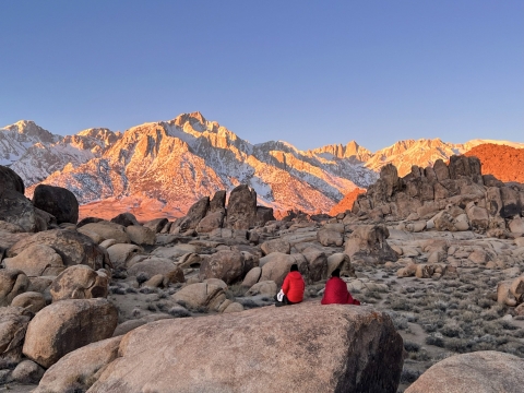 Two people sit on a large rock in the foreground of the picture, with their backs facing the camera as they stare off toward sunrise on the Eastern Sierrva Nevada.