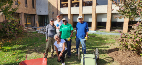 Five people stand in the yard of a building with wheelbarrows and other gardening tools