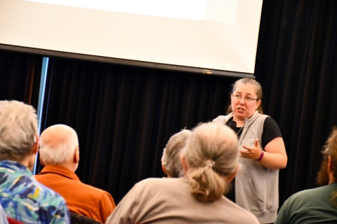 A woman speaks in front of a group of older people in front of a photo screen and a black curtain.