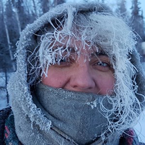 A person with frost-covered hair peeking out of a hood. Their mouth is covered by a neck gaiter and they smile with their eyes.