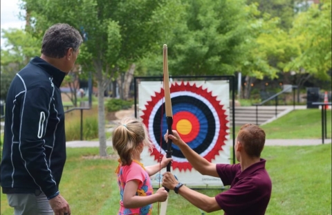 Park ranger helping child hold recurve bow with green grass, trees and target in background. 