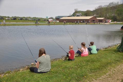 Four kids sitting on the grass, fishing a pond with people on the other side of the pond. 