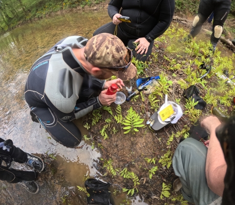 A man in a wetsuit is kneeling on a river bank. He is holding a freshwater mussel in his left hand, and is holding a squirt bottle in his right hand. Underneath the mussel is a plastic rubbermaid container. 