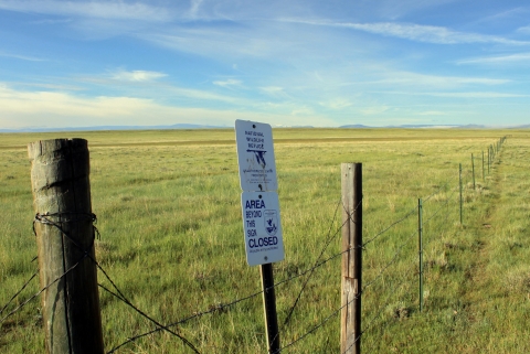 A border of the Mortenson Lake National Wildlife refuge is marked by a wire fence and signs. The field beyond the fence consists of green and yellow grasses and the sky is blue above with wispy white clouds.