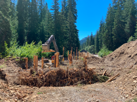 A worksite in a dry riverbed with heavy equipment and large wooden structures standing in the site. Tall green trees and blue sky in the background.