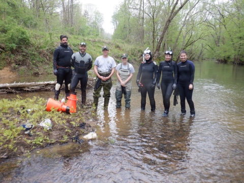 A group of seven people standing in a river. Five people are in wetsuits and two are in waders.