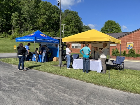 A blue USFWS canopy next to a yellow canopy. People are standing around the tables that are underneath the canopies. 