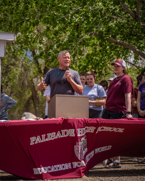 A man standing at a podium giving a speech while surrounded by other people. Green trees in background.