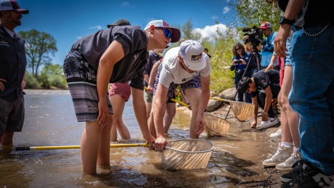 Students standing in a river while holding fishing nets above the water. On the shore, there is a group of people standing.