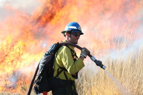 A man wearing yellow fire gear holding a hose tends to a prescribed fire