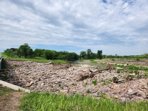 A rocky area extends from under a road crossing. It is disconnected from a river passing in the distance.