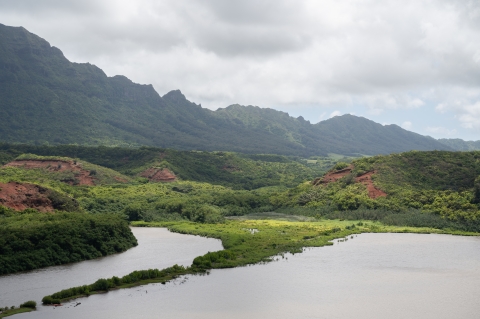 a stream cuts down a river valley to the left with a large pond on the right. Green jagged hills over look the valley to the left. It is a cloudy day. 