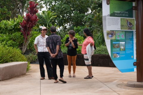 A group of four people stand in a paved walkway next to a sign about endangered wildlife. They are surrounded by lush greenery. Two of the people have huge smiles on their faces. They appear to be chatting. 