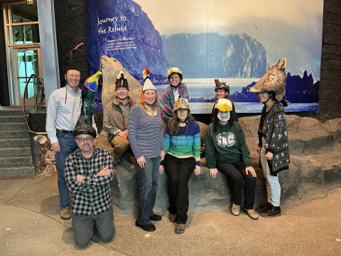 Nine people wear costume hats of different sea birds and one fox. They stand in front of a visitor center backdrop photo of ocean cliffs. They stand in a group to smile for a photo.